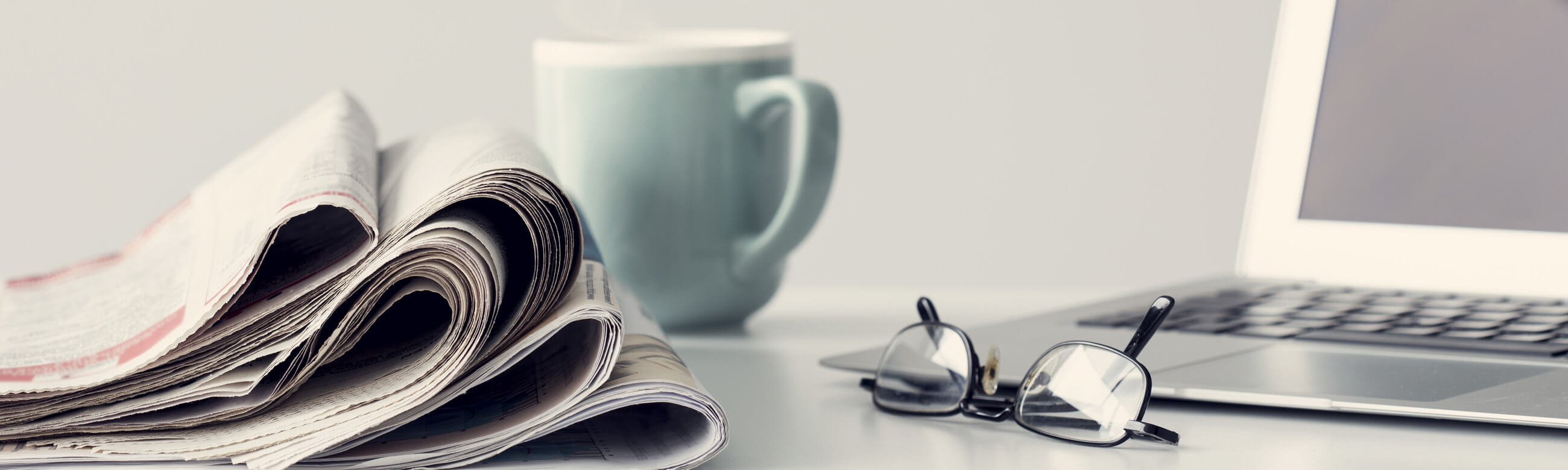 a stack of newspapers, eyeglasses, mug, and laptop atop a white table